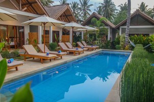 Un área junto a la piscina con sillones y sombrillas, rodeada de exuberante vegetación y palmeras.