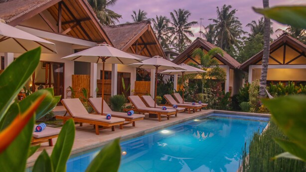 A poolside area with lounge chairs and umbrellas, surrounded by tropical plants and palm trees. - Cozy Cottages Lombok (Senggigi)