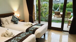 A hotel room with two beds, a nightstand with a lamp, and a view of a garden through a glass door. - Cozy Cottages Lombok (Senggigi)
