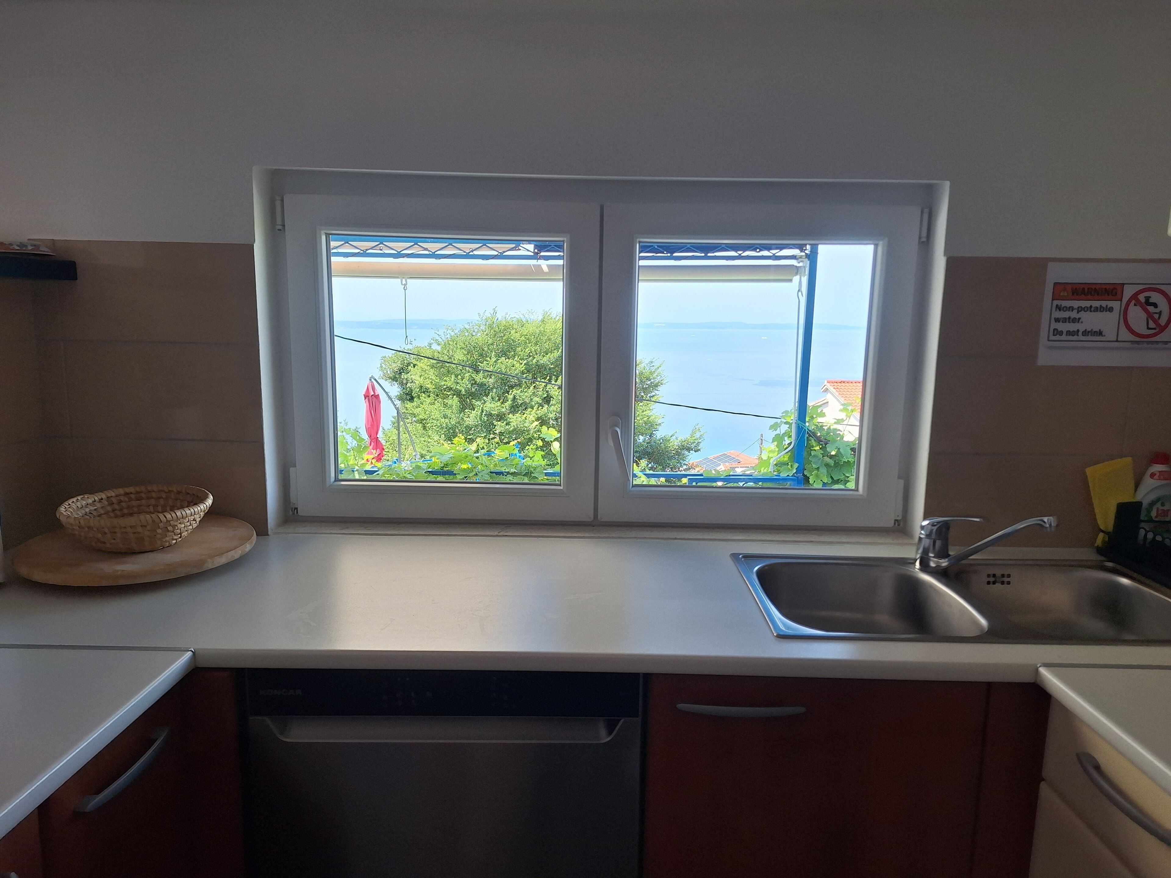 Une cuisine équipée d’un évier à deux bacs, d’un égouttoir et offrant une vue sur la mer par une fenêtre.