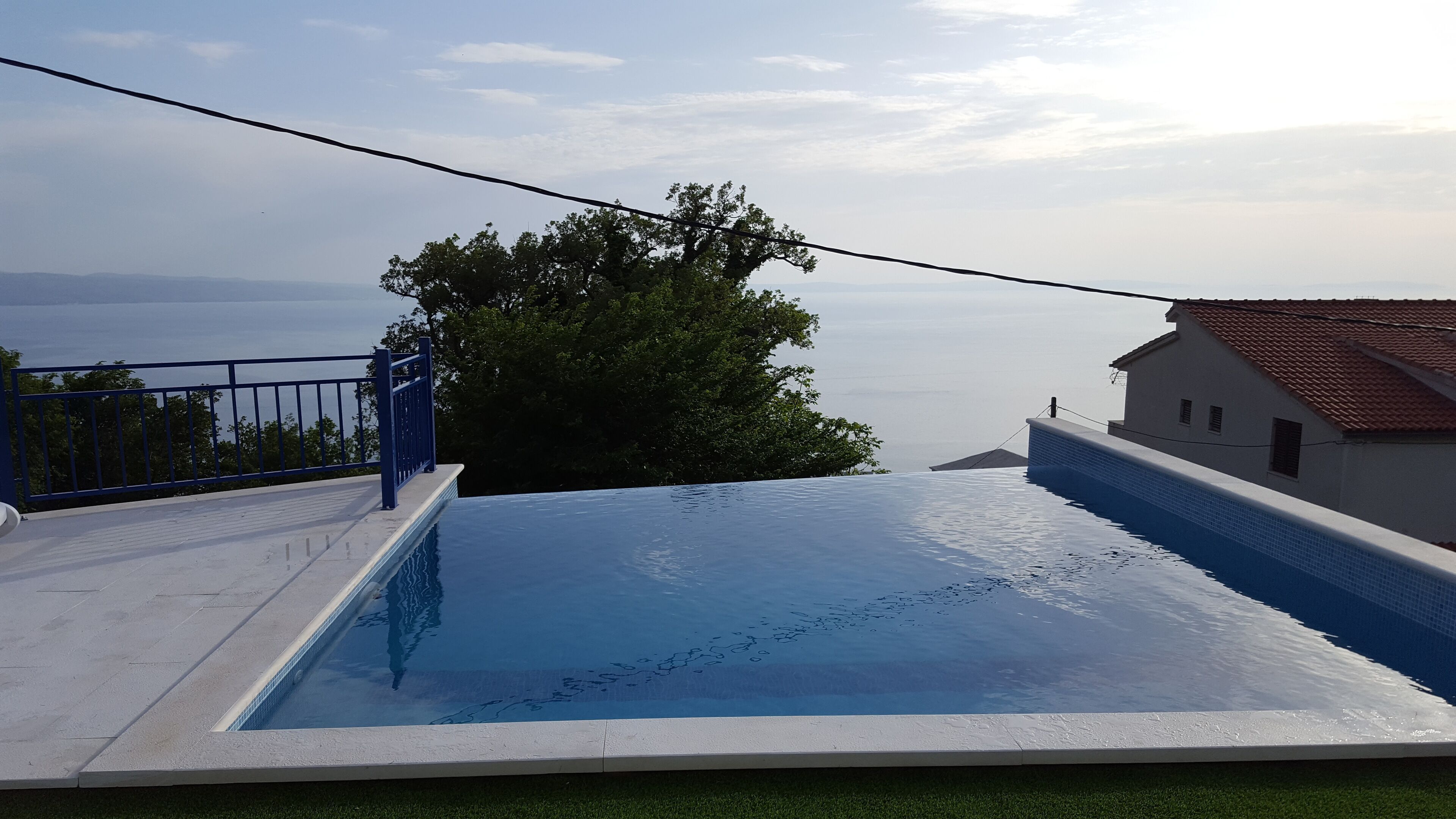 A swimming pool with a clear view of the sea in the background, a blue railing, and a white building with a red roof.