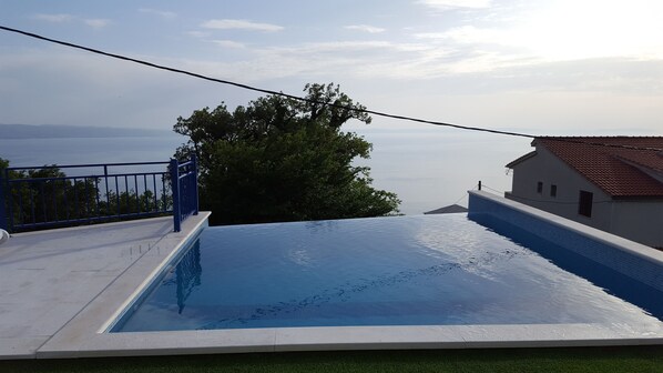 A swimming pool with a clear view of the sea in the background, a blue railing, and a white building with a red roof.