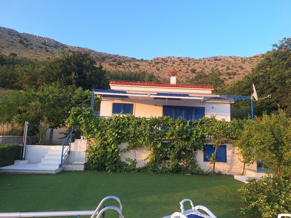 A modern two-story house with a white facade, blue shutters, and a red-tiled roof. It features a small staircase leading to the entrance, surrounded by lush greenery and a well-maintained lawn. - Villa Blue - spectacular view and heated pool, Split riviera, Croatia (Podstrana)