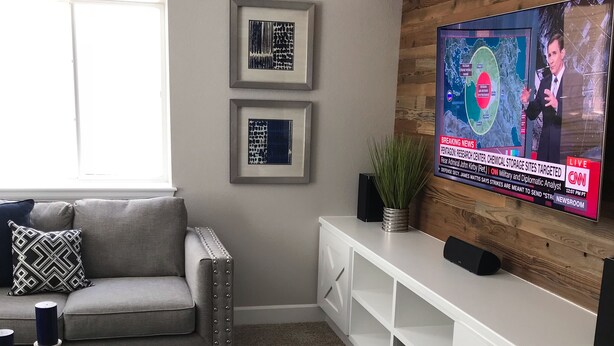 A modern living room with a grey sofa, a coffee table with decorative items, a TV mounted on a wooden wall displaying a news broadcast, and framed artwork on the wall. - A Fabulous home in the Rocky Mountain State (Centennial)