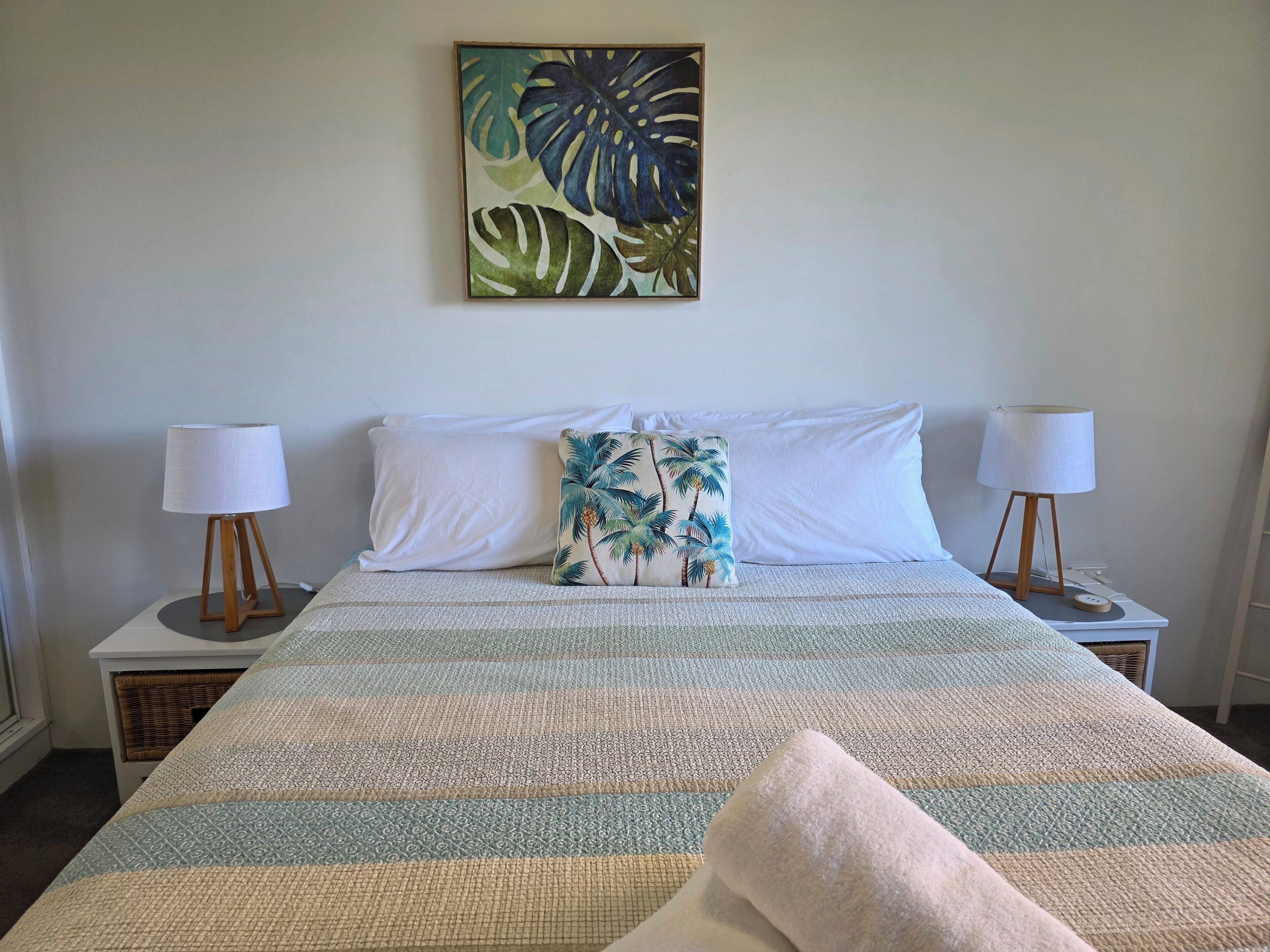A neatly made bed with a striped blanket, two white pillows, and a tropical-themed pillow. There are two bedside lamps on wooden stands.