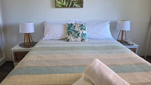 A neatly made bed with a striped blanket, two white pillows, and a tropical-themed pillow. There are two bedside lamps on wooden stands. - CHILL-OUT BEACHSIDE * coastal cool on Main Beach. Stroll to cafes, shops & lake! (Forster)