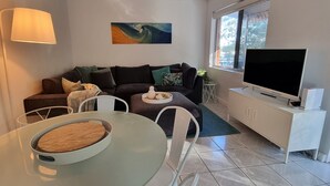 A living room with a sofa, a coffee table, a TV, and a dining table. - CHILL-OUT BEACHSIDE * coastal cool on Main Beach. Stroll to cafes, shops & lake! (Forster)