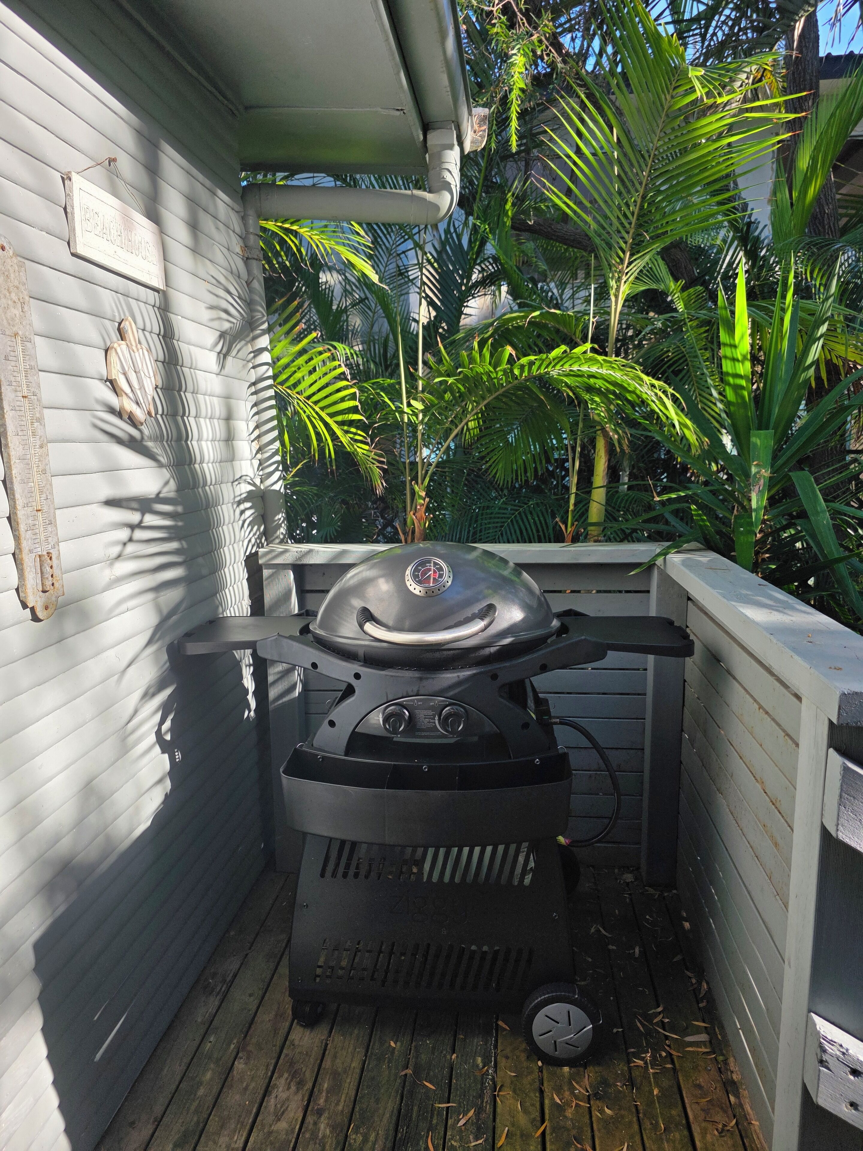 Un barbecue sur une terrasse en bois, avec une maison blanche en arrière-plan et des plantes tropicales tout autour.