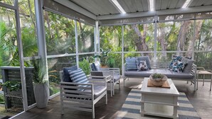 Une terrasse couverte avec un canapé, des fauteuils et une table basse, entourée de plantes en pot et de grandes fenêtres.