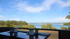Ein Balkon mit Tisch, zwei Blumentöpfen und Blick auf einen Fluss sowie weiter entfernte Bäume.