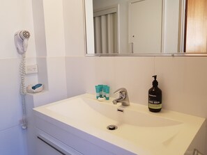 Kylpyhuone, jossa on valkoinen pesuallas, peili, hiustenkuivaaja ja hygieniatarvikkeita.