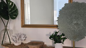 Ein weißes Konsolentische mit einem Spiegel, einer Glasflasche mit grünen Schilfhalmen, einer weißen Korallen-Skulptur, einer Holzschale und einer Topfpflanze.