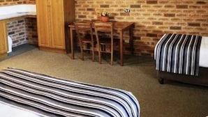 A room with a brick wall, a television mounted on the wall, a wooden desk with a chair, a bed with striped bedding, and a wardrobe.