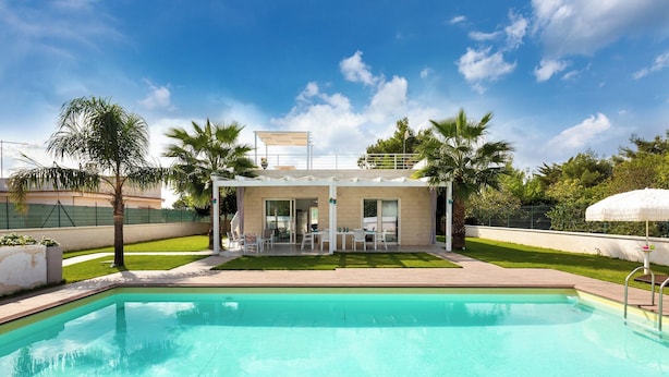 A swimming pool with a covered patio area, palm trees, and a clear sky.