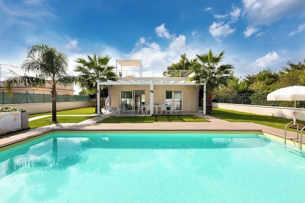 Ein Swimmingpool mit überdachter Terrasse, Palmen und blauem Himmel.