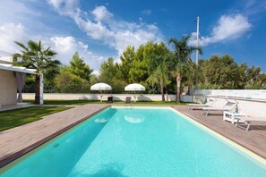 Ein rechteckiger Pool mit Holzterrasse, zwei weißen Sonnenschirmen und Liegestühlen.