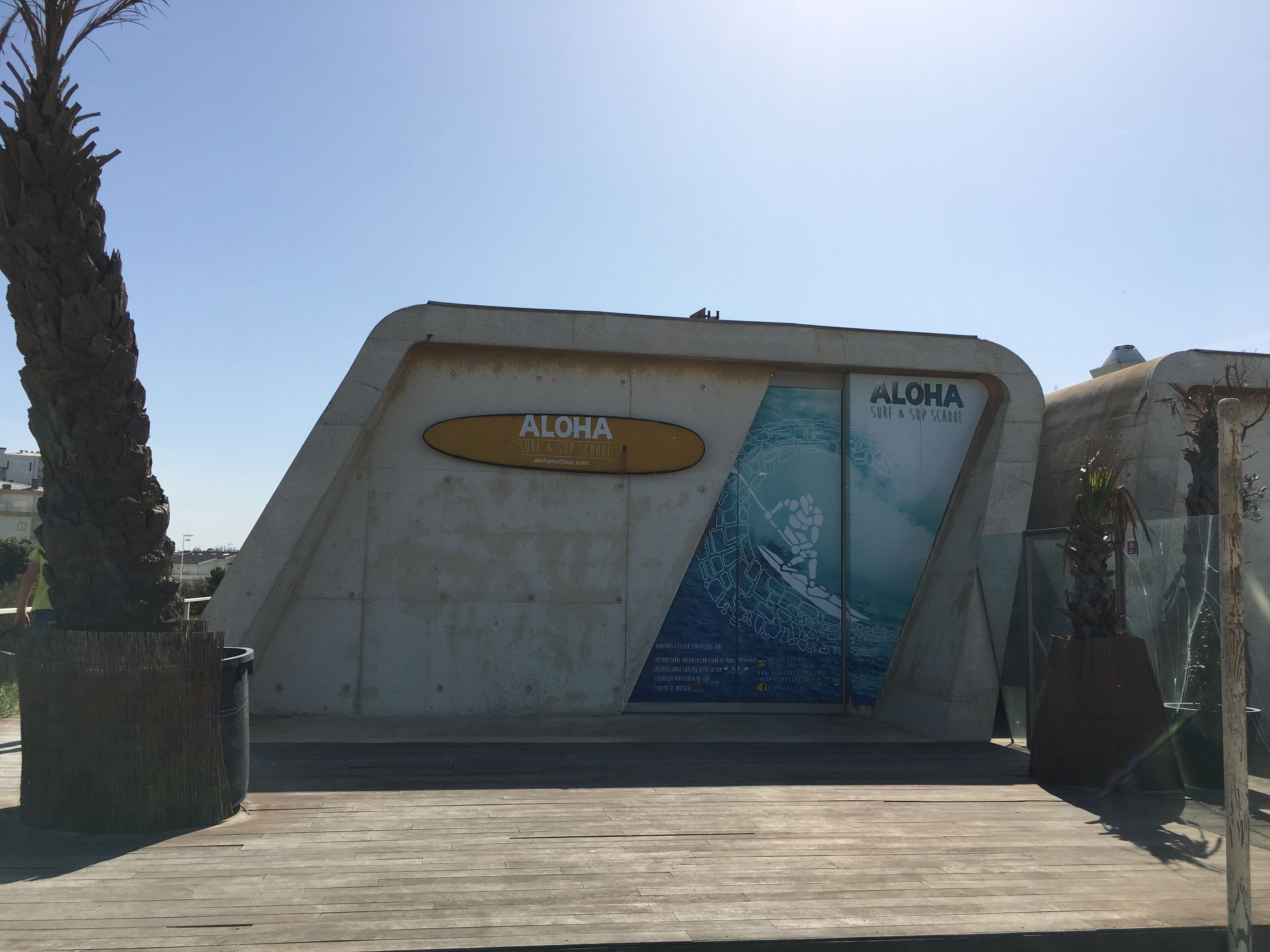 A concrete structure with a sign reading "ALOHA SURF & SUP SCHOOL" and a poster of a surfer.