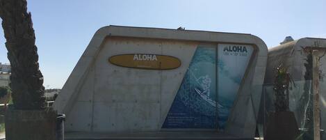 A concrete structure with a sign reading "ALOHA SURF & SUP SCHOOL" and a poster of a surfer.
