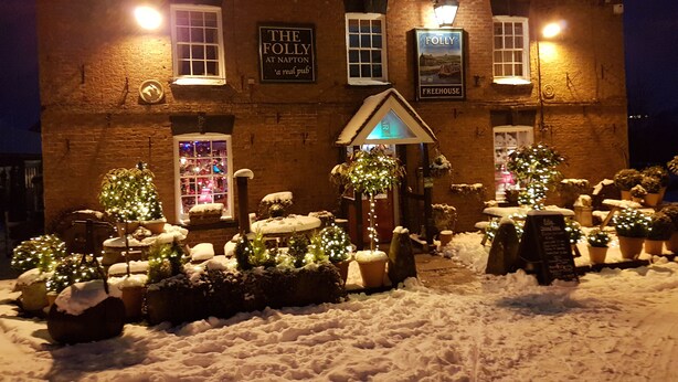 A brick building with illuminated signs reading "The Folly at Napton" and "Freehouse." Snow-covered ground and potted plants are visible.