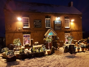 A brick building with illuminated signs reading "The Folly at Napton" and "Freehouse." Snow-covered ground and potted plants are visible.