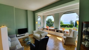 A spacious living room with a large TV, comfortable seating, and a view of the sea through arched windows.