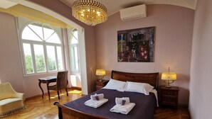 A spacious bedroom with a large bed, two bedside tables with lamps, a wooden floor, a chair, and a large window with curtains.