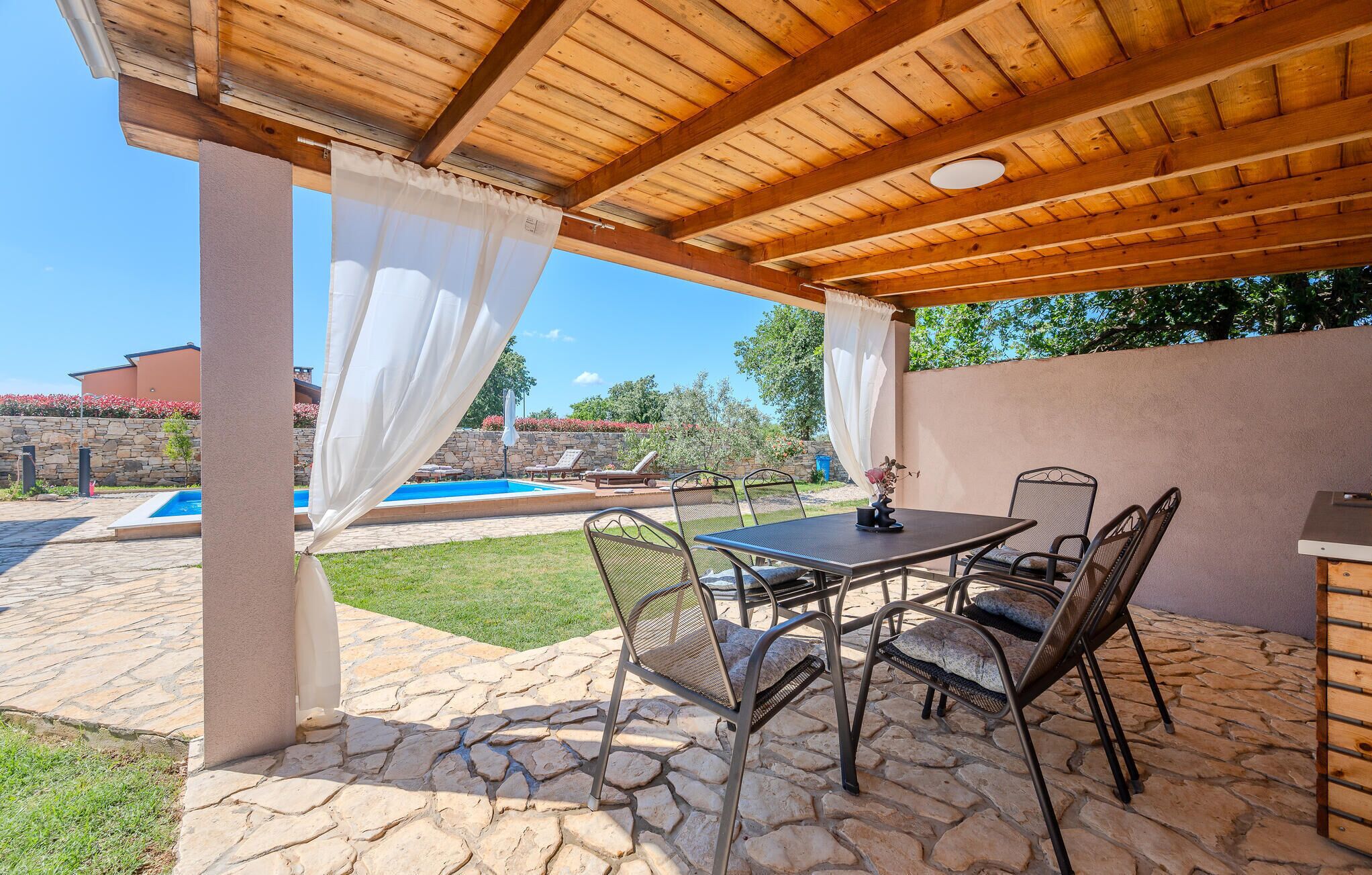 An outdoor patio with stone flooring, a table with chairs, and a pool in the background.