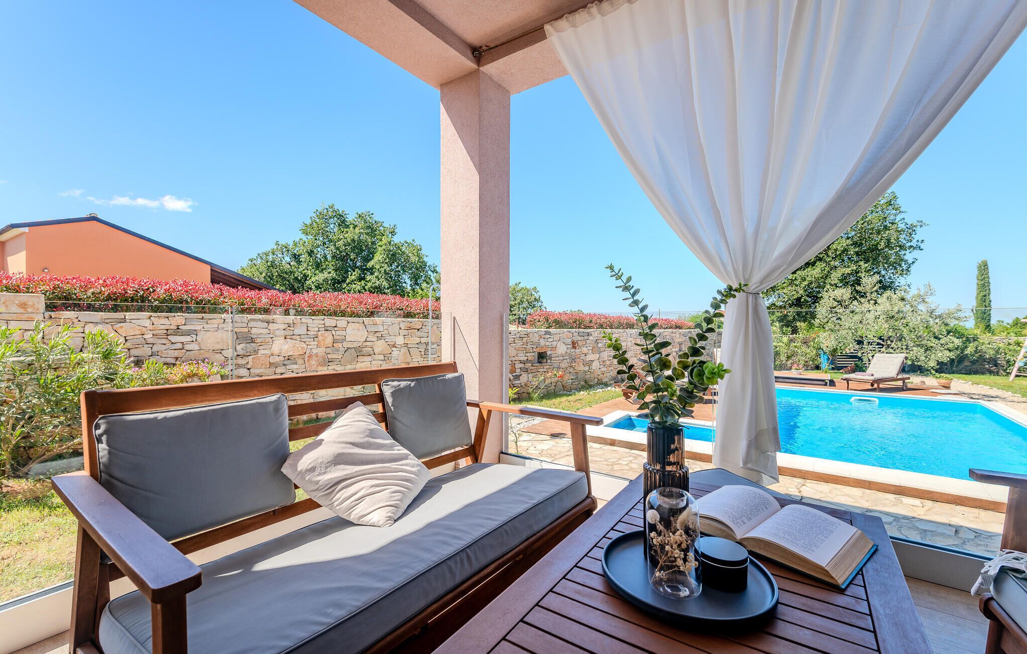 A covered outdoor seating area with a pool view, stone wall, and greenery.