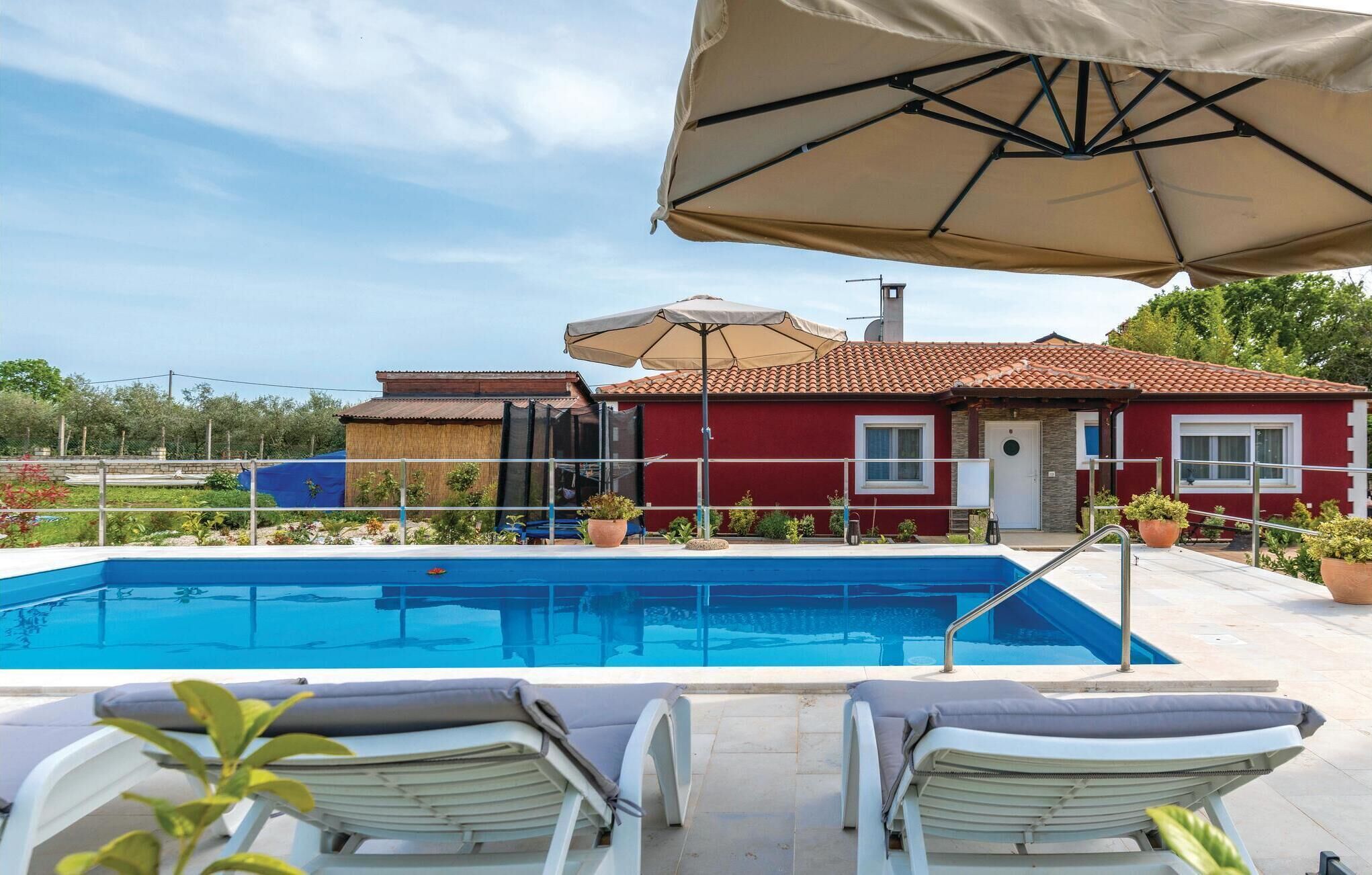 A swimming pool with lounge chairs and umbrellas, a red building with a tiled roof, and a clear sky.