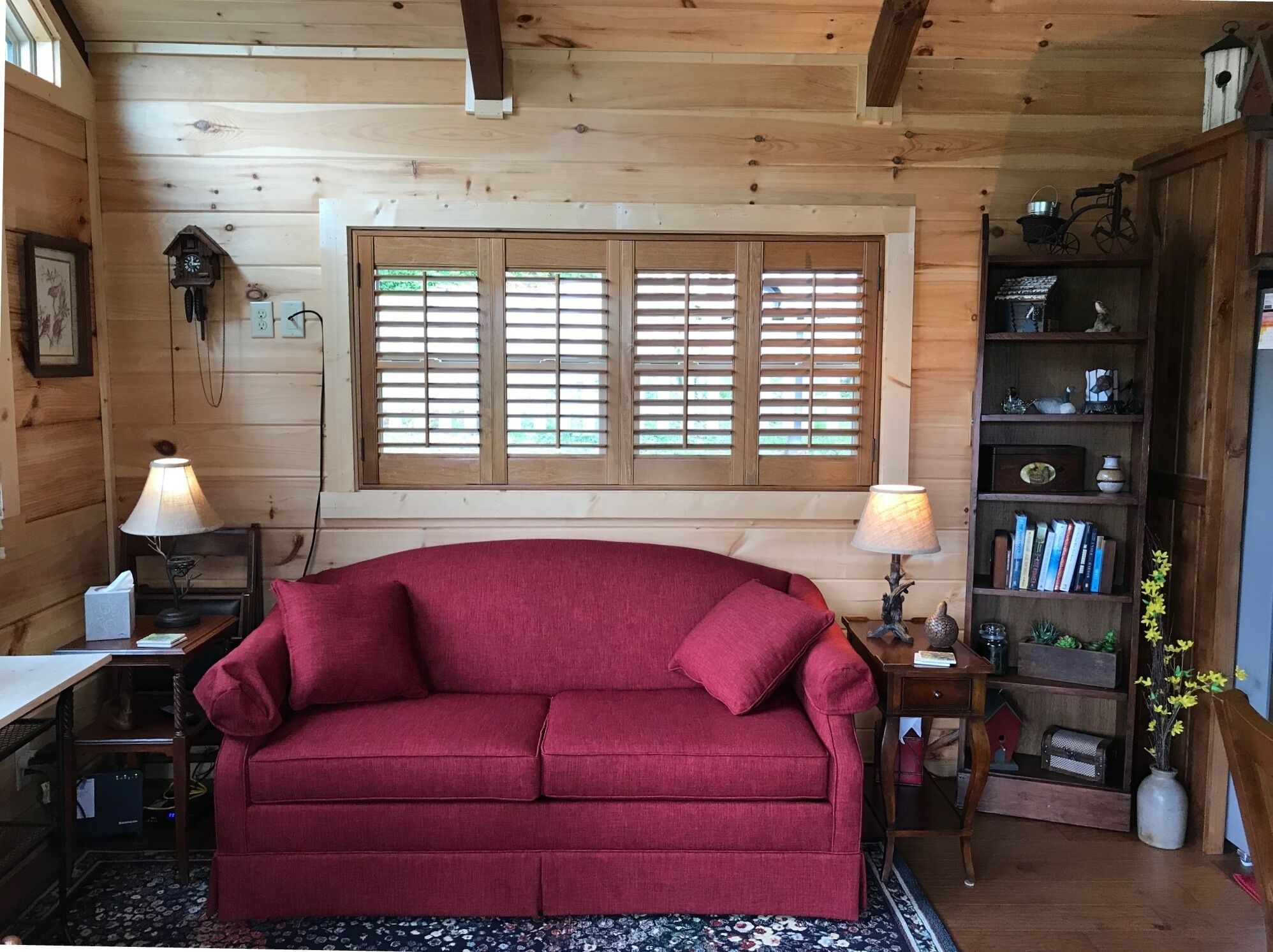 Una habitación acogedora con un sofá rojo, paredes de madera, una ventana con contraventanas, una estantería y una lámpara.