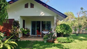 Una casa con veranda, un tavolo da pranzo e sedie. Si apre su un giardino con diverse piante e un prato.