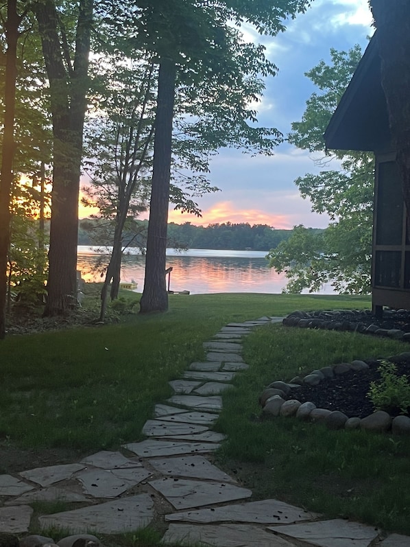 A stone pathway leading to a lake with trees on either side and a building in the background.