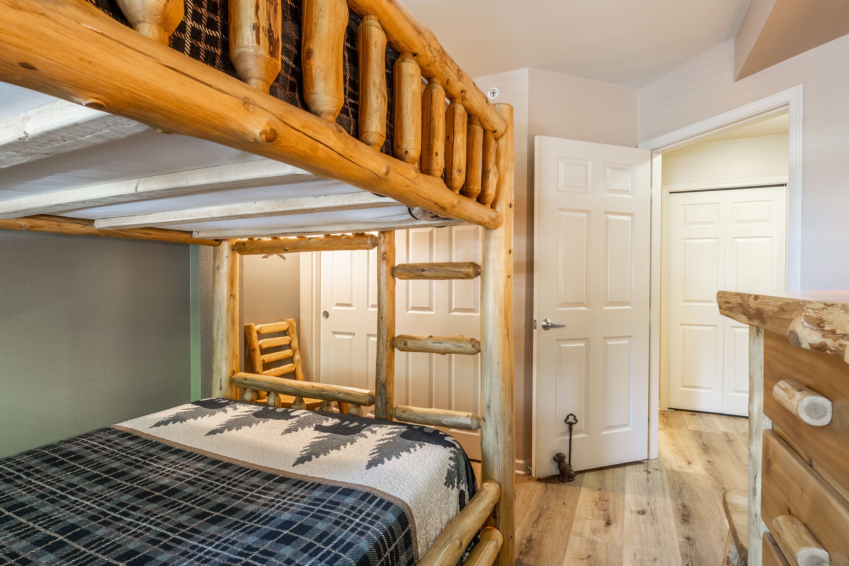 Uma cama beliche de madeira rústica com escada, uma cadeira e uma porta em um quarto com piso de madeira.
