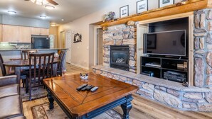 A living room with a stone fireplace, a television, and a dining area with a table and chairs. - Mammoth Green #105 - Nearly Ski-in Ski-Out Updated 2 Bedroom 2 Bath, Sleeps 6 (Mammoth Lakes)