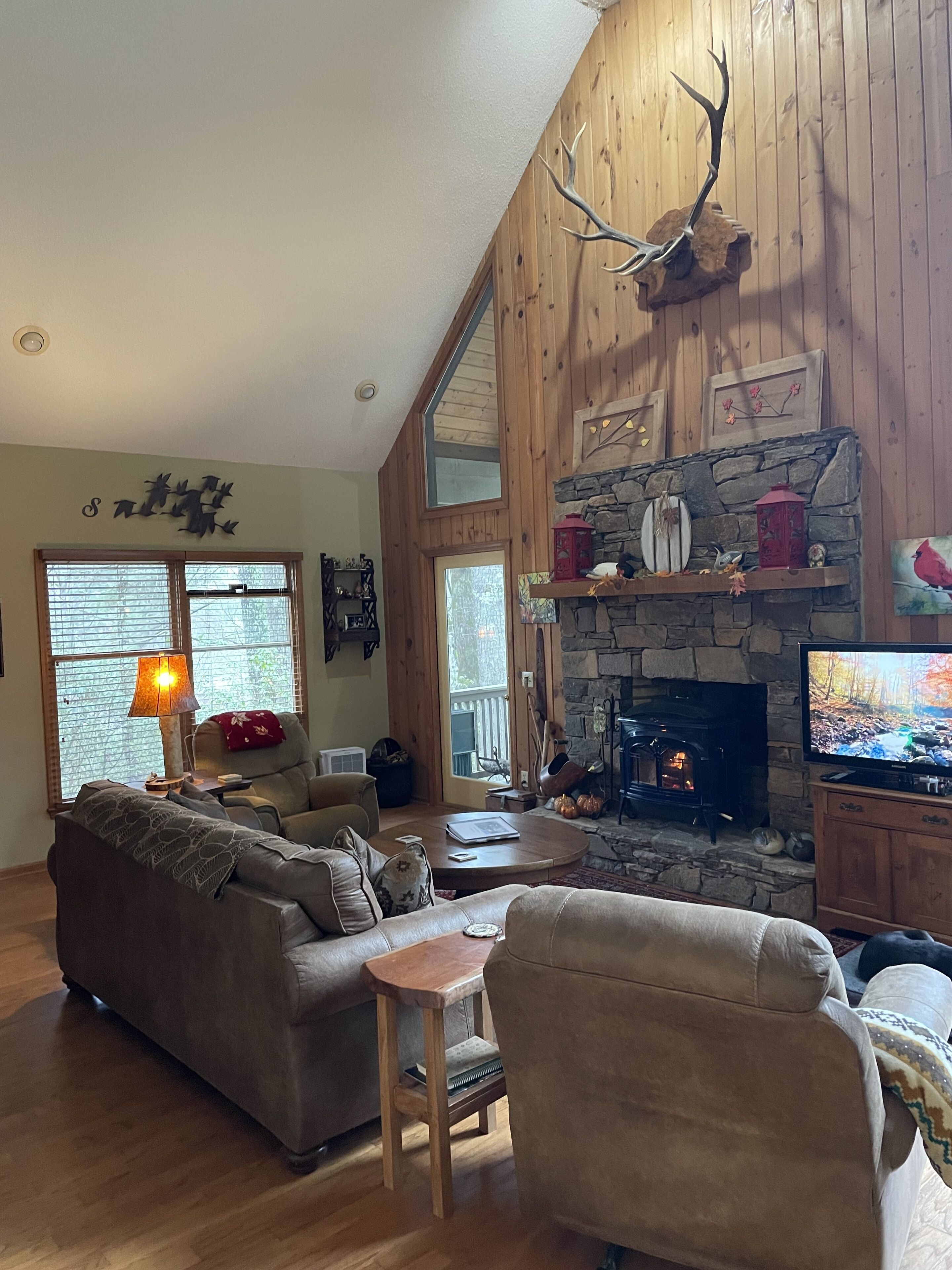 A cozy living room with a stone fireplace, a large window, and a wooden ceiling.