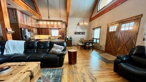 Un vaste espace de vie comprenant un canapé en cuir noir, une table basse en bois et un tapis. Au fond, on aperçoit une cuisine avec des meubles en bois et un luminaire suspendu.