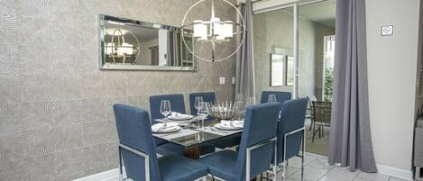 A dining area with a glass table, blue chairs, a chandelier, and a large mirror on the wall.