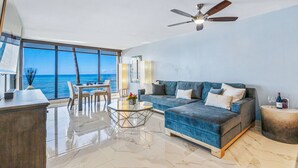 Ein geräumiges Wohnzimmer mit großem Fenster und Meerblick, Deckenventilator, blaues Sofa und Glastisch.