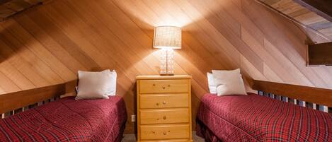 A small, cozy attic bedroom with two beds, a wooden dresser, and a lamp.