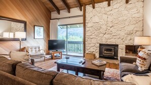 A cozy living room with a stone wall, wooden ceiling beams, a fireplace, and a large flat-screen TV. - Snowcreek #97 - Great Views. Large. 2 bedroom + loft, 2 bathroom, 3-level condo. Sleeps 8 (Mammoth Lakes)