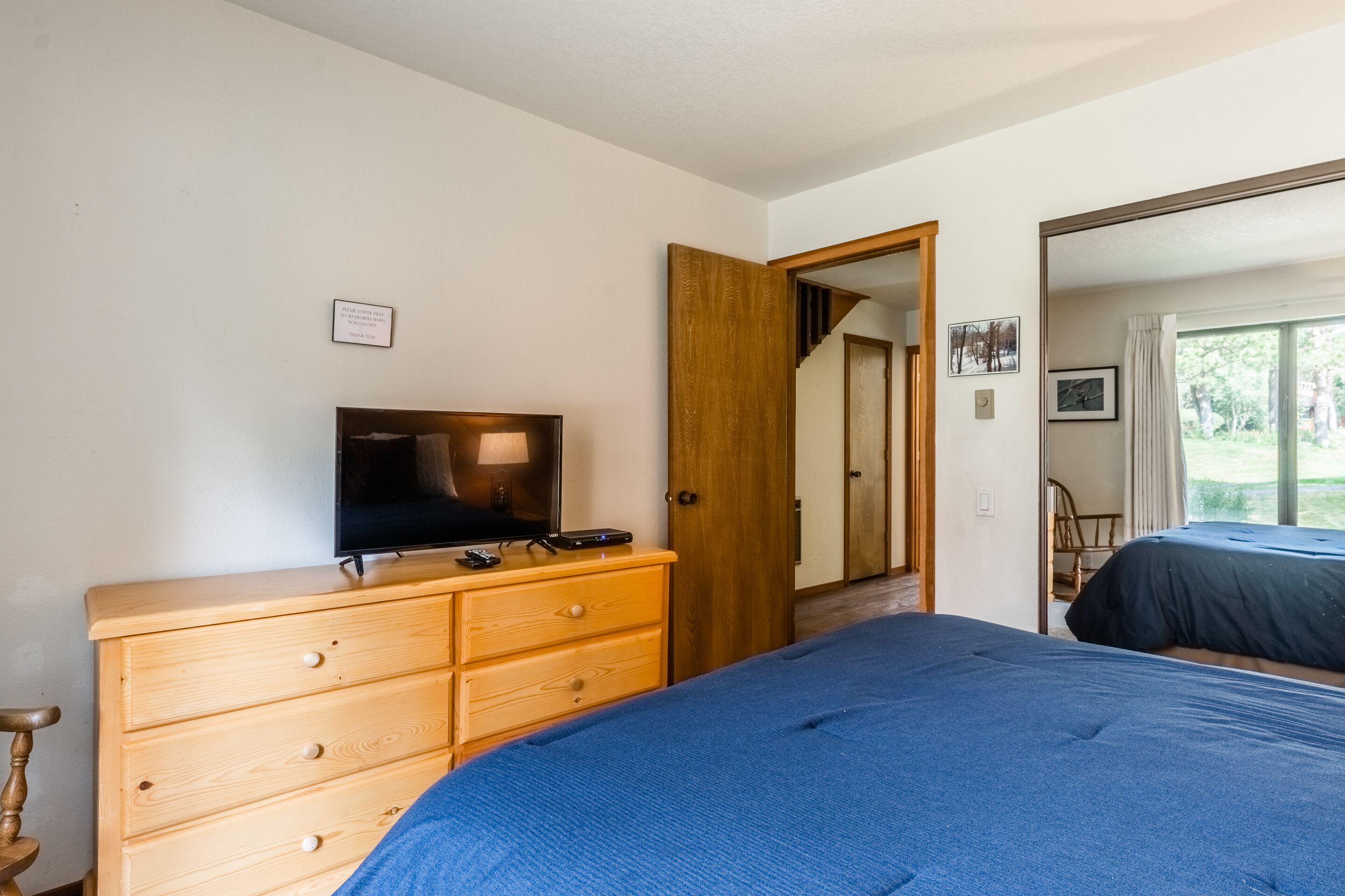 A bedroom with a bed, a wooden dresser, a television, and a door leading to another room.