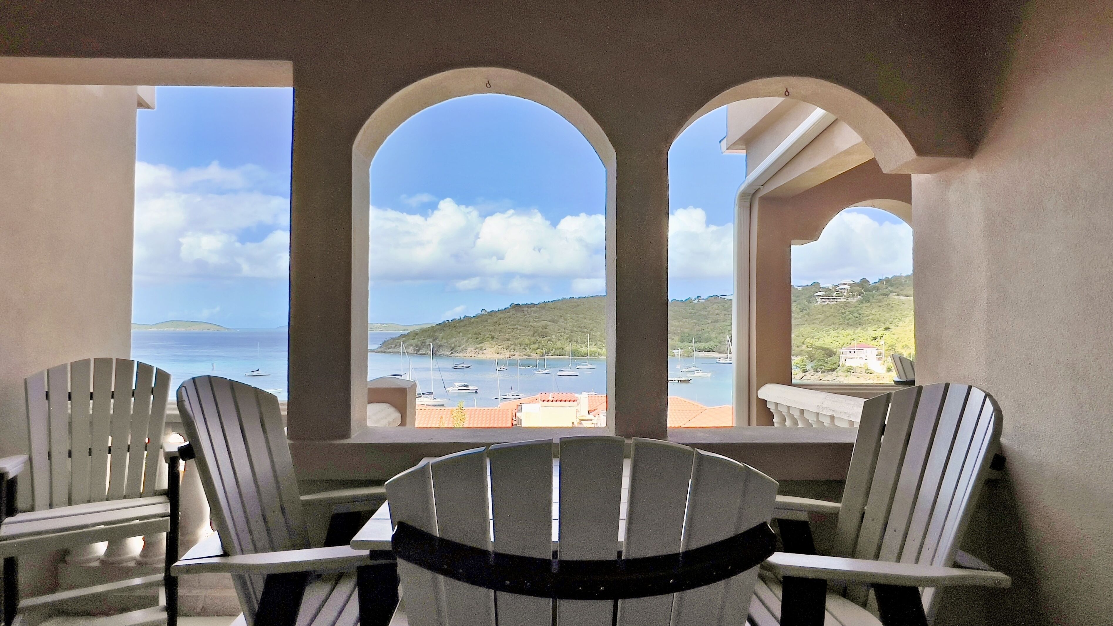 Uma varanda com três cadeiras Adirondack, uma mesa e vista para o mar e as colinas.