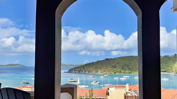 Blick auf die Küste durch ein Rundbogenfenster, mit Booten im Hafen, einer hügeligen Landschaft und blauem Himmel.