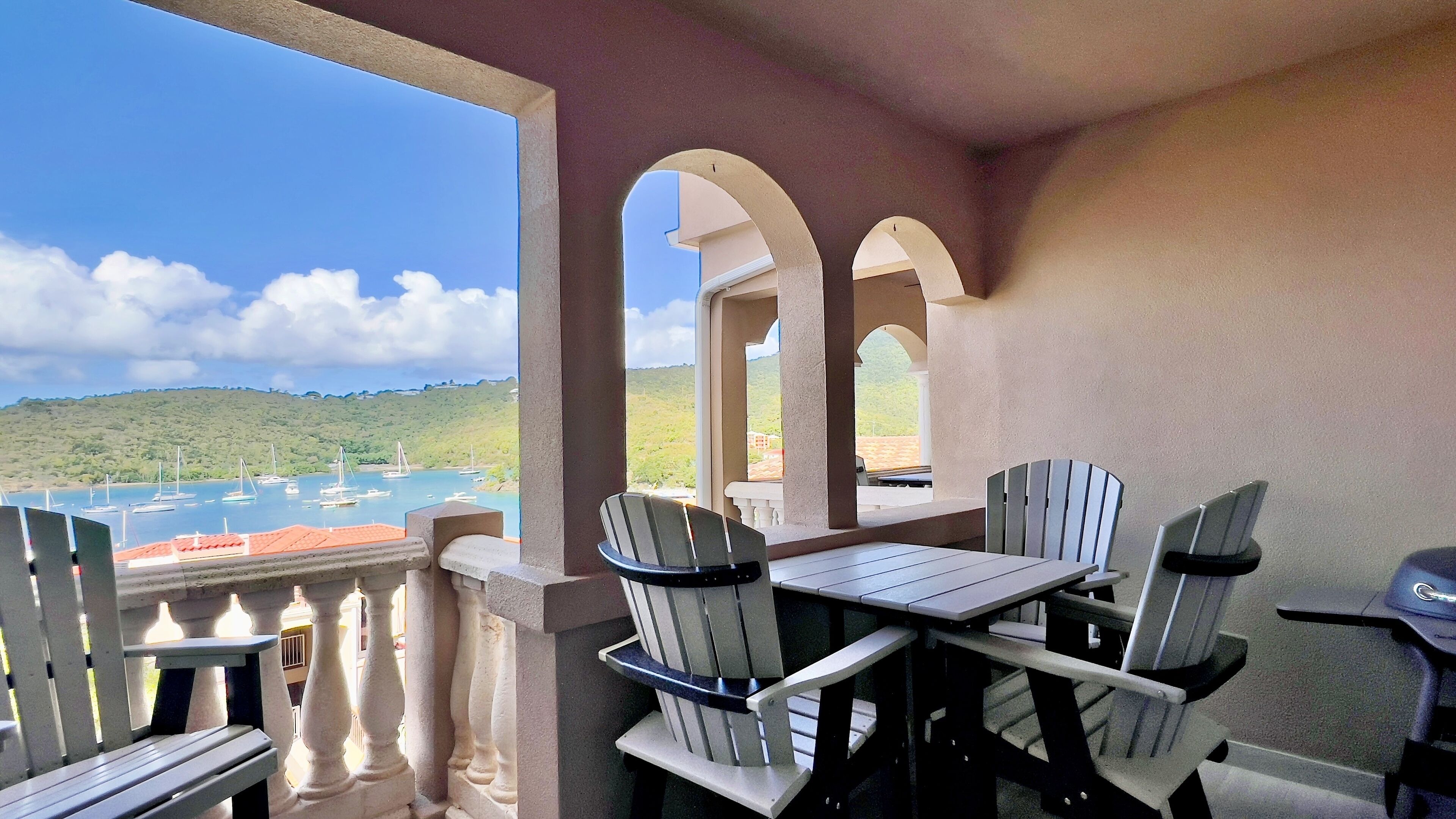 Ein Balkon mit Tisch und Stühlen, der auf einen Yachthafen mit Booten und eine hügelige Landschaft blickt.