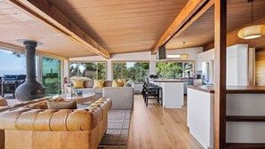 A modern living room with a large leather sofa, a kitchen with white cabinets, and a large window overlooking a garden. - Newly Renovated Oceanfront Retreat in Bolinas with Spa and huge fenced yard  (Bolinas)