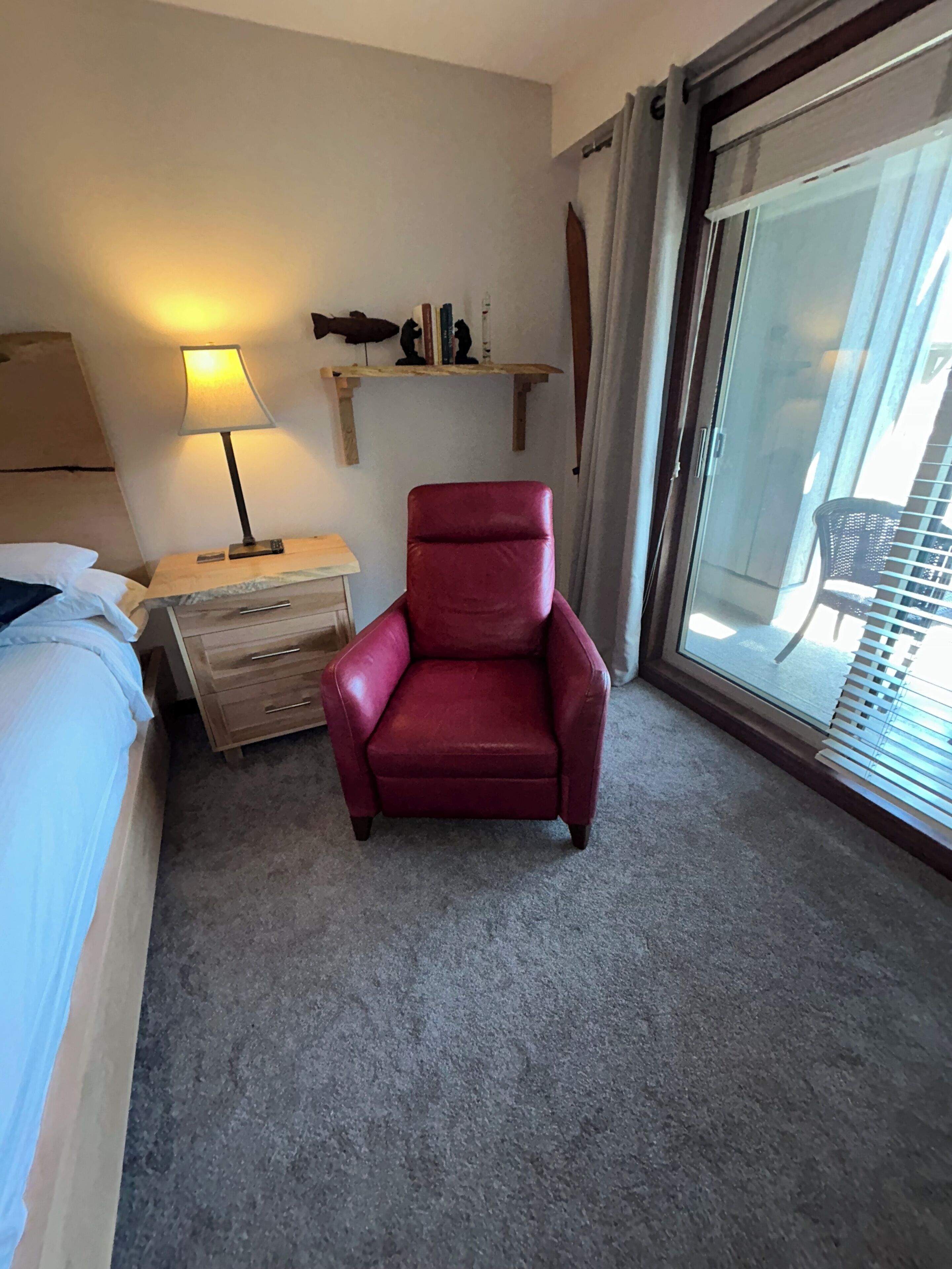 A hotel room with a bed, a red armchair, a nightstand with a lamp, a small shelf with decorative items, and a sliding glass door leading to a balcony.
