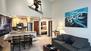 A modern living room with a grey sofa, a coffee table, and a TV. A kitchen is visible in the background with wooden cabinets and a microwave. - XL Luxury Penthouse. Top 1%. The Aspens. A/C, Pool, 3 hot tubs, ski-in ski-out.  (Whistler)