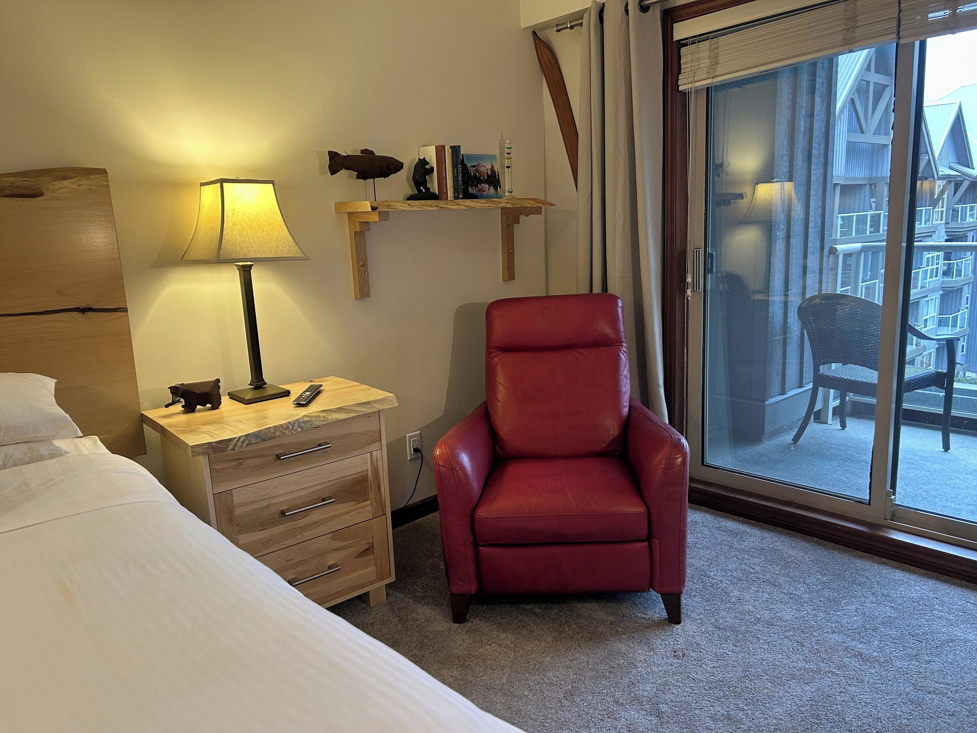 A hotel room with a bed, a red leather chair, a wooden nightstand with a lamp, a small shelf with books and a fish decoration, and a sliding glass door leading to an outdoor area.