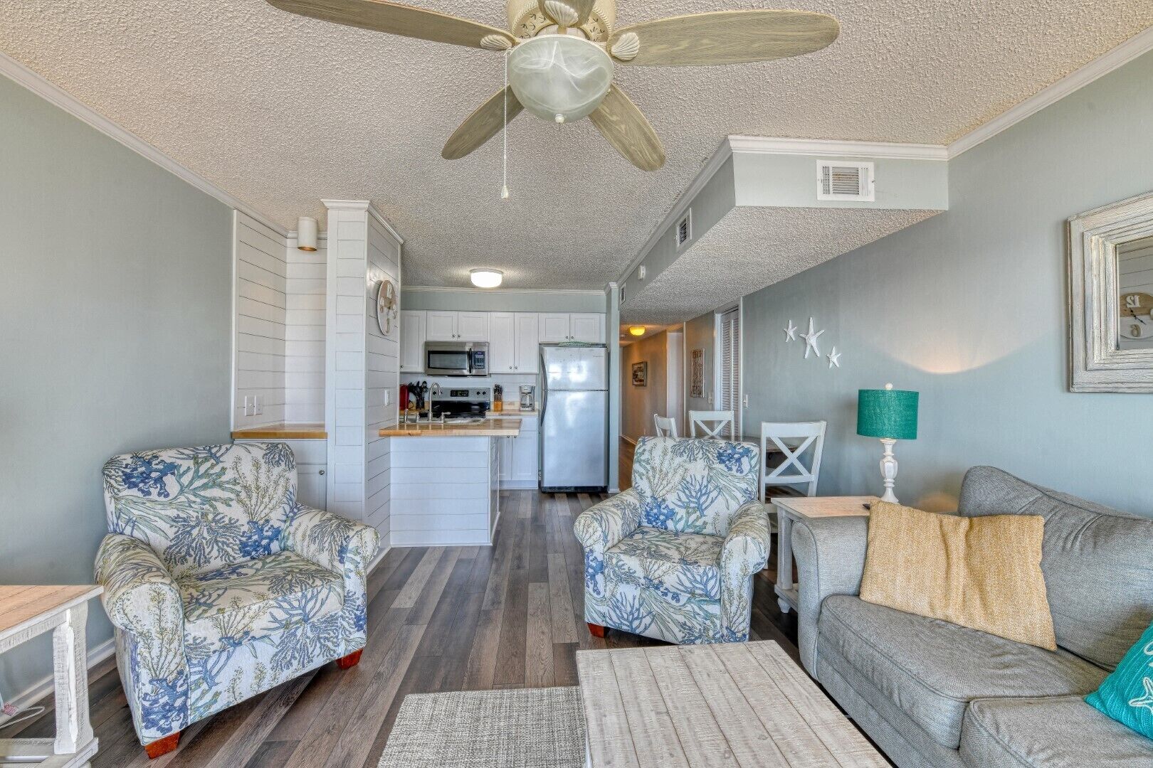 A compact living space with a kitchenette, two patterned armchairs, a grey sofa, and a ceiling fan.