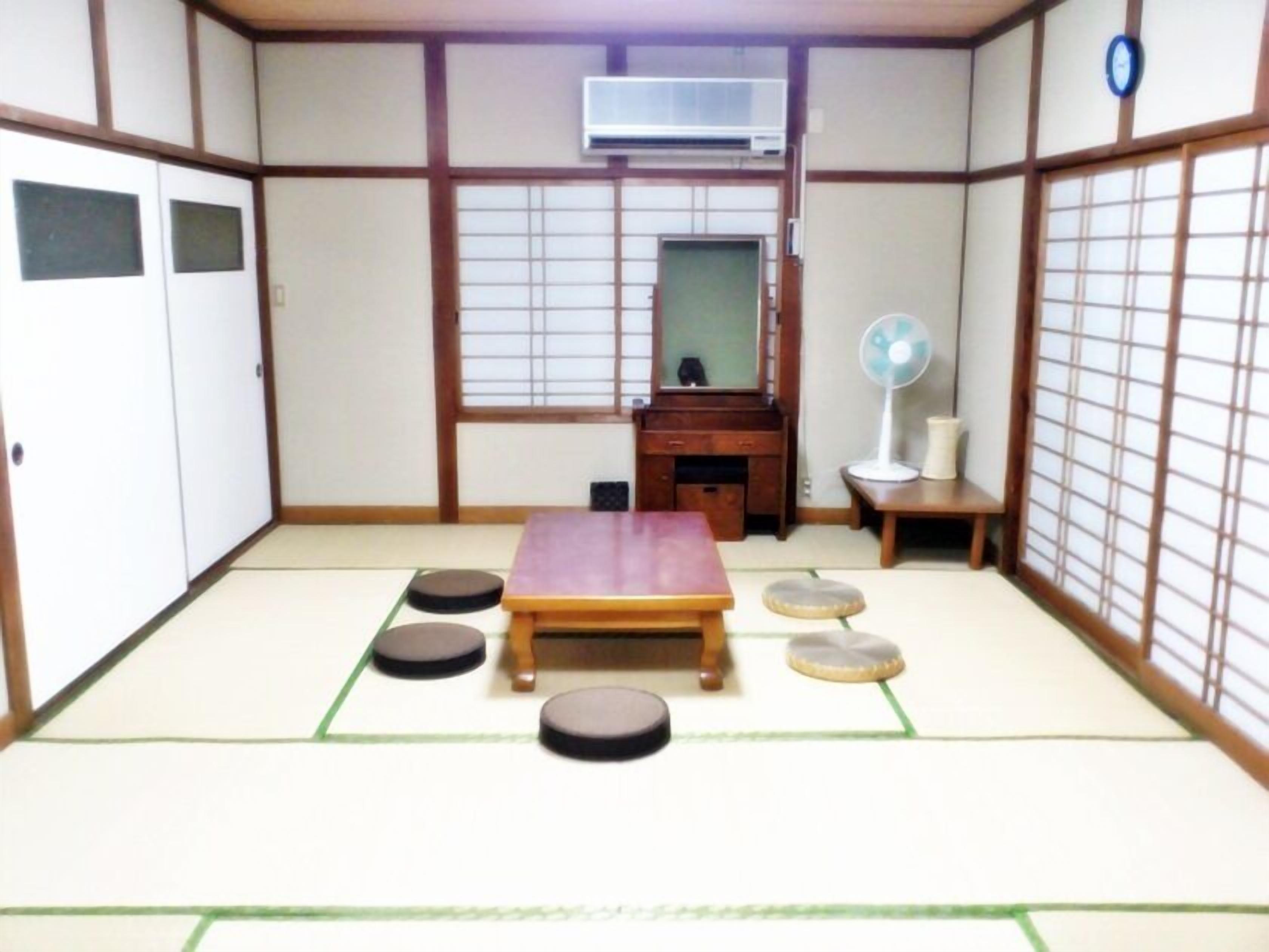 Een kamer met tatami matten, een futonbed, een houten bureau, een spiegel, een ventilator en een schuifdeur.